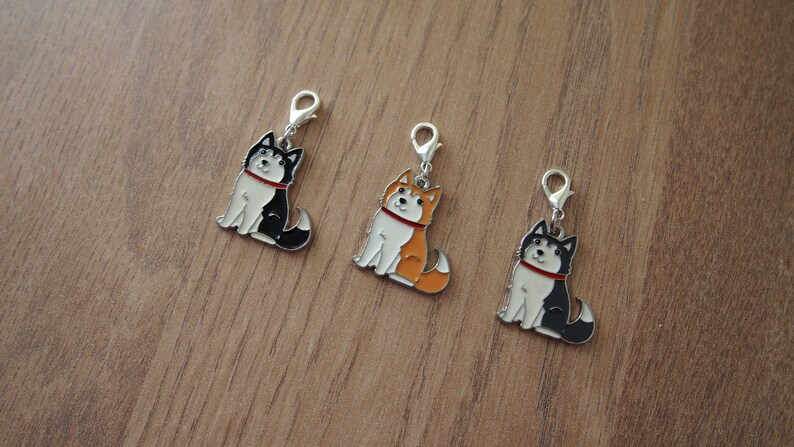 May include: Three metal charms featuring cartoon husky dogs in different colors: black and white, orange and white, and black and white. The charms have a silver finish and a loop for attaching to a keychain or bag.