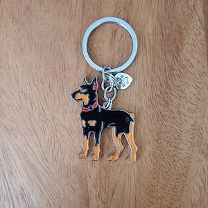May include: A black and orange Doberman Pinscher keyring with a silver ring and a small charm. The dog has a red collar and is standing. The keyring is on a wooden surface.