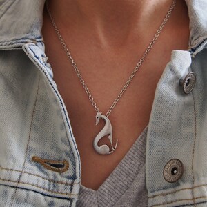 May include: Silver-toned pendant necklace featuring a stylized swan design. The pendant hangs from a delicate chain, creating a minimalist and elegant aesthetic. The necklace is worn over a gray top and a light blue denim jacket.