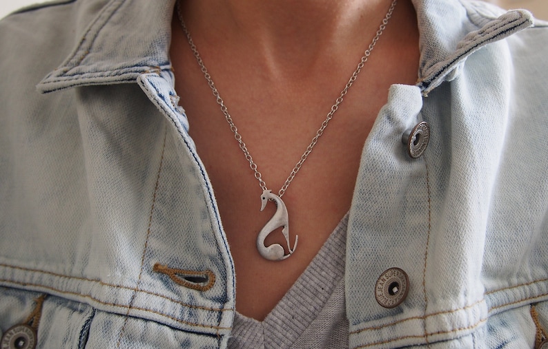 May include: Silver-toned pendant necklace featuring a stylized swan design. The pendant hangs from a delicate chain, creating a minimalist and elegant aesthetic. The necklace is worn over a gray top and a light blue denim jacket.