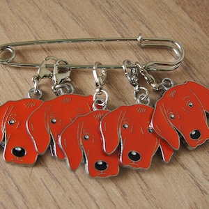 May include: A silver safety pin with six red dog charms attached. The charms are all the same design, featuring a red dog's head with black eyes and nose.