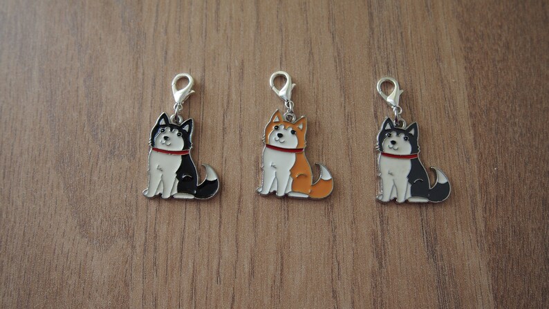 May include: Three metal charms featuring cartoon husky dogs in black, orange, and white. Each charm has a silver loop for attaching to a bag or keychain.