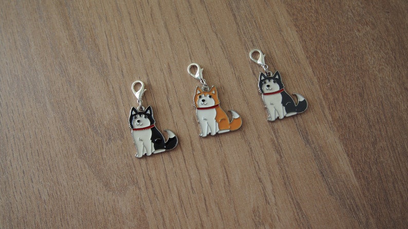 May include: Three silver charm pendants featuring cartoon husky dogs. The dogs are sitting and have different colored fur: black and white, orange and white, and black and white with a red collar.