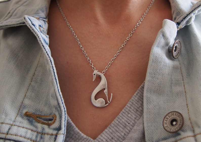 May include: A silver-colored pendant necklace featuring a stylized greyhound dog design. The dog pendant hangs from a delicate silver chain. The necklace is worn over a gray sweater and a light blue denim jacket.