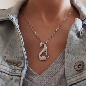 May include: Silver-toned pendant necklace featuring a stylized greyhound dog design. The pendant hangs from a delicate chain. The person is wearing a light blue denim jacket over a grey shirt.