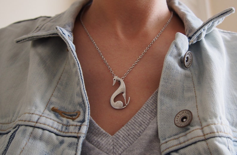 May include: Silver-toned pendant necklace featuring a stylized greyhound dog design. The pendant hangs from a delicate chain. The person is wearing a light blue denim jacket over a grey shirt.