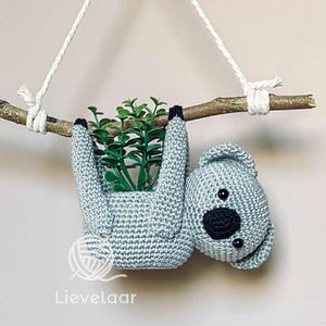 Può includere: Un koala di uncinetto grigio appeso a un ramo con una piccola pianta in vaso. Il koala tiene la pianta con le braccia e ha occhi neri e un naso nero.