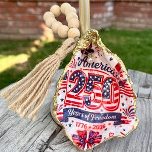 May include: A decorative oyster shell ornament with gold-painted edges, featuring a patriotic design with the text America 250 Years of Freedom 1776-2026, accented by a small pearl bead, a jute tassel, and a loop of natural wooden beads for hanging.