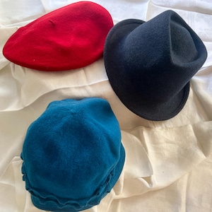 May include: Three hats in different colors rest on a white cloth. A red beret, a dark blue fedora, and a teal cloche hat are displayed. The hats are made of a soft material and are likely for fashion or costume purposes.