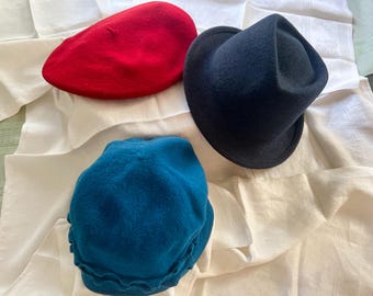 May include: Three hats in different colors rest on a white cloth. A red beret, a dark blue fedora, and a teal cloche hat are displayed. The hats are made of a soft material and are likely for fashion or costume purposes.