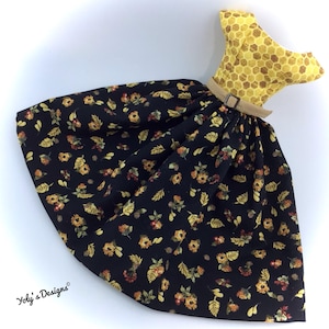 Puede incluir: Un objeto decorativo con forma de vestido. La parte superior es amarilla con un patrón de panal, y la falda es negra con estampados florales y de hojas amarillas, naranjas y marrones. Un cinturón beige con hebilla define la cintura. El artículo es decorativo.