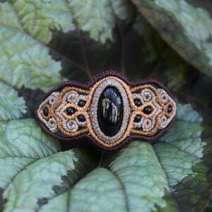 May include: A black gemstone is set in a brown and beige macrame bracelet. The bracelet is made with intricate knotting and features a geometric design.