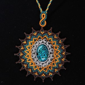 Puede incluir: Un collar de macramé hecho a mano con una piedra central ovalada de turquesa. El colgante presenta intrincados patrones en marrón, verde azulado, dorado y gris. El collar tiene un cordón a juego con un nudo decorativo.
