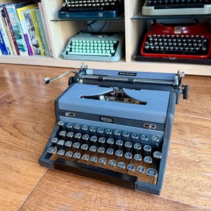 May include: A vintage Royal typewriter in a gray and black color scheme, with a full keyboard of round keys. The typewriter sits on a wooden surface, with other typewriters visible on shelves in the background.