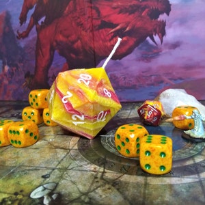 Puede incluir: Un conjunto de dados naranja y verde con puntos negros, una gran vela d20 amarilla y roja, un dado d20 rojo y una figura en miniatura en un tablero de juego.
