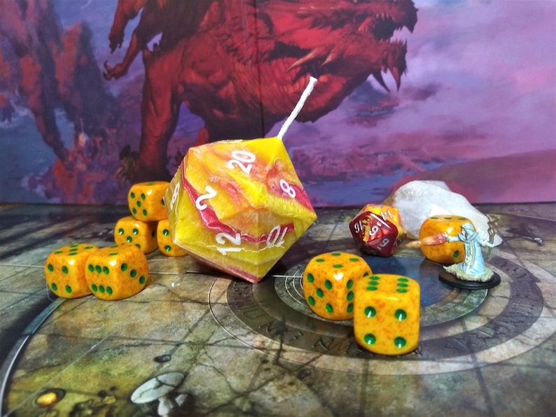 Puede incluir: Un conjunto de dados naranja y verde con puntos negros, una gran vela d20 amarilla y roja, un dado d20 rojo y una figura en miniatura en un tablero de juego.