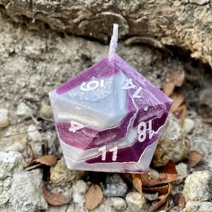 May include: A purple and white candle shaped like a 12-sided die with the numbers 1, 4, 7, 8, 10, and 11 visible.