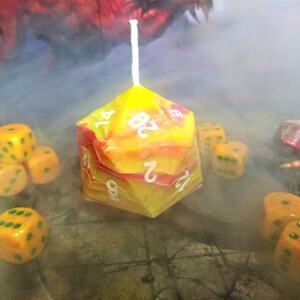 Puede incluir: Una vela d20 amarilla y roja con n&uacute;meros blancos. La vela est&aacute; rodeada de dados amarillos y verdes sobre una superficie de madera. Hay humo en el fondo.