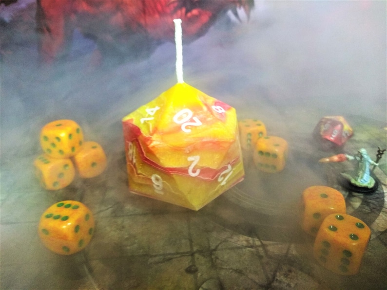 Puede incluir: Una vela d20 amarilla y roja con n&uacute;meros blancos. La vela est&aacute; rodeada de dados amarillos y verdes sobre una superficie de madera. Hay humo en el fondo.