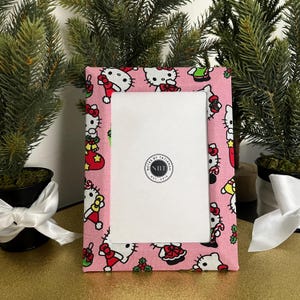 May include: Pink picture frame with a white mat and a Hello Kitty Christmas design. The frame has a circular logo that says "Notes by Tatiana" and "NBT" with "Est. 2018" underneath.