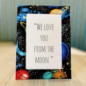 May include: A fabric-covered picture frame with a black background featuring a colorful space-themed pattern of planets, stars, and galaxies. The frame holds a white card with the text "WE LOVE YOU FROM THE MOON."