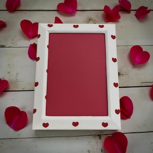 May include: A white picture frame with a red interior and small red hearts around the border. Scattered around the frame are red heart-shaped petals, creating a romantic and decorative theme.