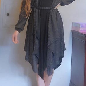 Könnte beinhalten: Ein schwarzes Kleid mit einem schwarz-weiß gemusterten Cardigan. Der Cardigan hat einen langen, fließenden Schnitt und ist in der Taille mit einem Gürtel versehen.