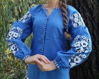 Puede incluir: Una blusa azul bordada con motivos florales blancos en las mangas. La blusa tiene cuello en V y mangas largas.