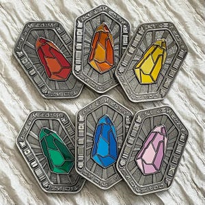 Peut inclure: Six broches hexagonales en métal, chacune présentant une forme cristalline centrale en rouge, orange, jaune, vert, bleu ou rose. Les cadres argentés ont un fond texturé avec des lignes rayonnantes et des symboles décoratifs sur la bordure.