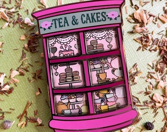 May include: A pink enamel pin shaped like a shopfront with the text "TEA & CAKES" above a display of teacups, cakes, and pastries. The pin has a detailed design with pink, white, and black accents.