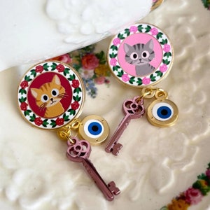 May include: Two gold-tone cat charm earrings with a red and pink floral design. Each earring features a cat with a different coloured fur, a gold key charm, and a blue evil eye charm.