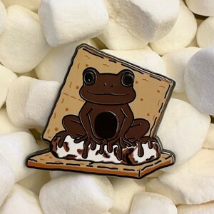May include: A brown frog sitting on a graham cracker with chocolate and a white marshmallow. The frog is wearing a smile and has large eyes. The graham cracker is surrounded by white marshmallows.