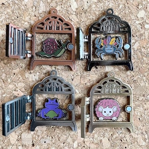 May include: Four enamel pins featuring different animals inside a cage. The cages are all the same design, but in different colours. The animals are a snail, a crab, a frog, and a sheep.