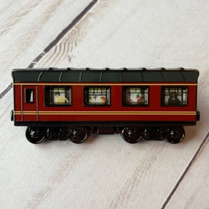 May include: A red and black train carriage enamel pin with four windows showing passengers inside. The train carriage has a black roof and black wheels.