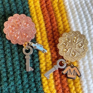Könnte beinhalten: Zwei goldfarbene Anstecknadeln mit einem Schneeflockenmuster. Die Nadel links ist roségoldfarben und hat einen kleinen Auto-Anhänger. Die Nadel rechts hat einen kleinen Pullover-Anhänger.