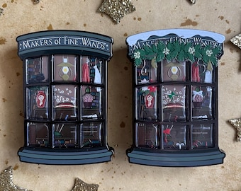 May include: Two enamel pins depicting a storefront window with the text "Makers of Fine Wands". The left pin shows the storefront in its regular state, while the right pin shows the storefront decorated for the holidays with a snowy roof, garland, and holly.