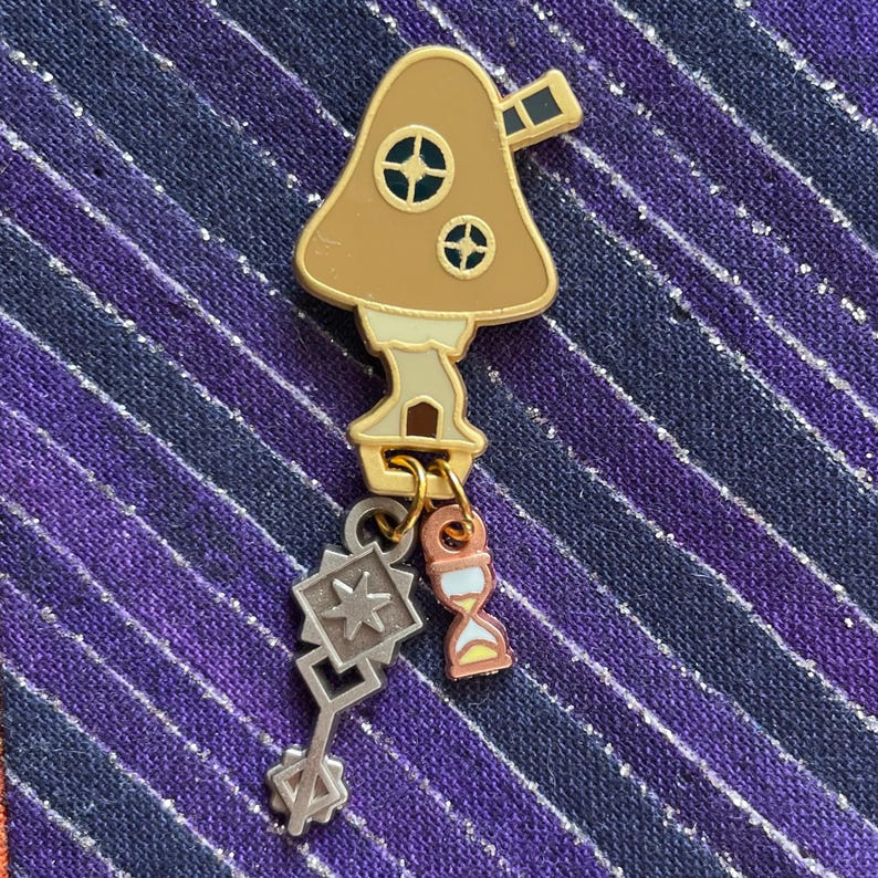 May include: A mushroom-shaped enamel pin with a gold border, featuring two circular windows and a small chimney. The pin has dangling charms, including a silver geometric shape and a small hourglass. The color palette includes brown, gold, and silver.
