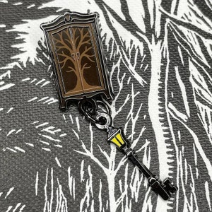 May include: A black metal pin shaped like a wardrobe with a brown tree inside and a key with a yellow light at the end.