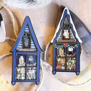 Op de afbeelding: Twee blauwe emaille pin-ornamenten in de vorm van huizen met besneeuwde daken. Beide huizen hebben een bord met de tekst "Post Office". De ramen zijn gevuld met feestelijke details, waaronder uilen, cadeaus en een sneeuwpop.