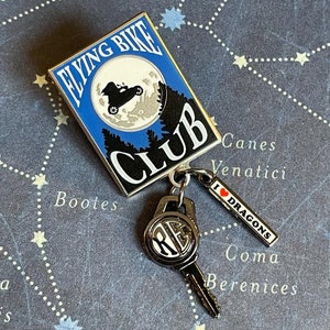 May include: A silver enamel pin featuring a flying bike in front of a blue moon with the text "Flying Bike Club". A silver key charm with the initials "RE" is attached to the pin with a silver chain. A red and white charm with the text "I ♥ Dragons" is also attached to the pin.