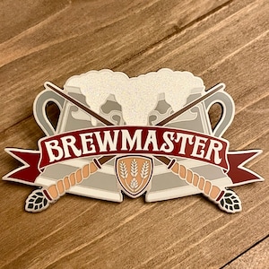 May include: A silver and red enamel pin with a beer mug design. The pin features two crossed wands, a banner with the text "BREWMASTER", and a hop leaf design.