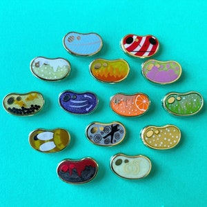 Puede incluir: Una colección de coloridos broches de esmalte con forma de gominola y bordes dorados. Los broches presentan varios diseños, como una pluma, llamas y una rodaja de naranja. El fondo es de color turquesa liso.