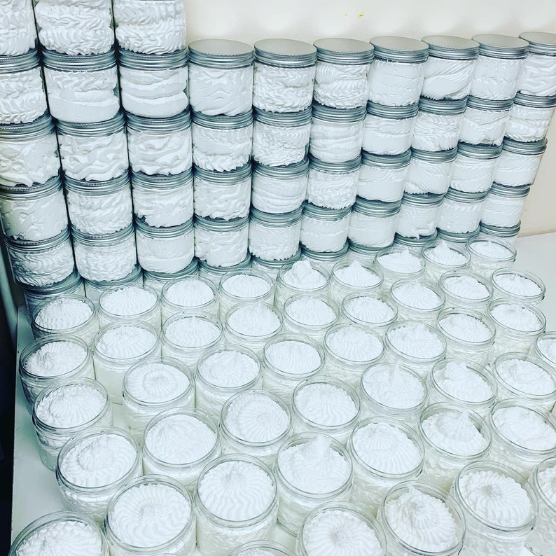 May include: A large collection of white whipped body butter in clear glass jars with silver lids. The jars are arranged in rows and columns, creating a visually appealing display.