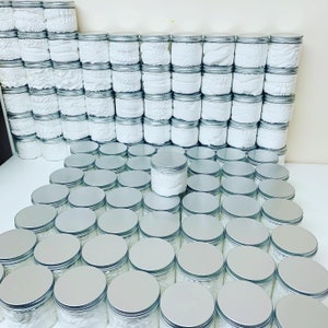 May include: A large collection of silver-lidded jars filled with white cream. The jars are arranged in rows on a white surface.