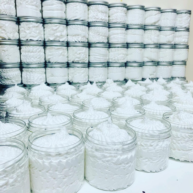 May include: A large collection of white whipped body butter in clear plastic jars with silver lids. The jars are arranged in rows and columns, creating a visually appealing display.