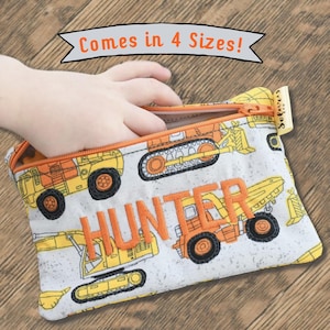 Op de afbeelding: Een rechthoekig stoffen etui met rits en een patroon van gele en oranje bouwvoertuigen op een lichtgrijze achtergrond. De naam HUNTER is in oranje draad op de voorzijde geborduurd. Een banner erboven vermeldt Comes in 4 Sizes!