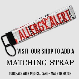 Op de afbeelding: Een witte stoffen polsband-sleutelhanger met een ruitpatroon en de in rood geborduurde tekst ALLERGY ALERT met een hartsymbool. De band is bevestigd aan een zwarte metalen sluiting en een ronde zwarte metalen sleutelring voor bevestiging.