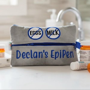 Op de afbeelding: Een grijs gewatteerd tasje met rits, voorzien van blauw borduurwerk met de tekst "Declan's EpiPen" en twee ronde iconen met blauwe doorgestreepte tekst "EGGS" en "MILK". Het tasje heeft een blauwe rits en een klein wit label aan de zijkant.