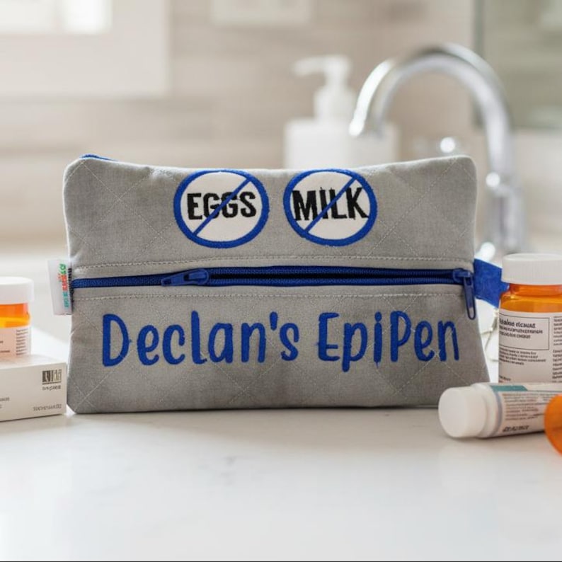 Op de afbeelding: Een grijs gewatteerd tasje met rits, voorzien van blauw borduurwerk met de tekst "Declan's EpiPen" en twee ronde iconen met blauwe doorgestreepte tekst "EGGS" en "MILK". Het tasje heeft een blauwe rits en een klein wit label aan de zijkant.