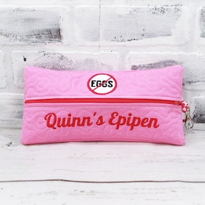 May include: Pink quilted zippered pouch with a red zipper and a red circle with a diagonal line through it and the word "EGGS" inside. The pouch also has the words "Quinn's Epipen" embroidered in red.